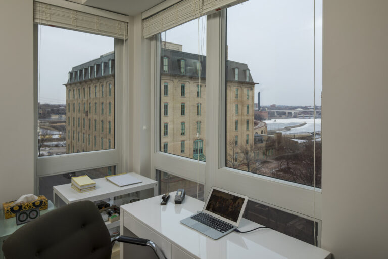 A modern office desk by large windows with a laptop, office supplies, and city buildings outside.