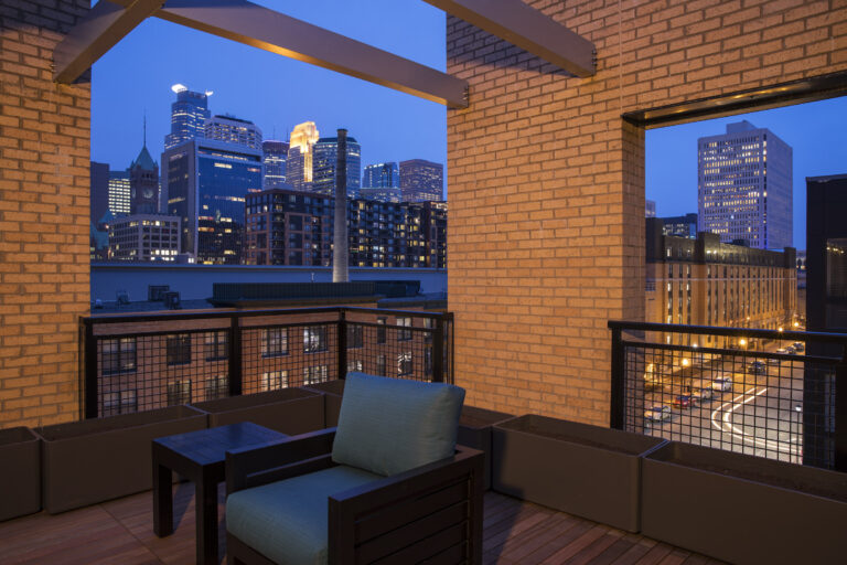 Rooftop patio with a chair, city skyline and tall buildings lit up at dusk.