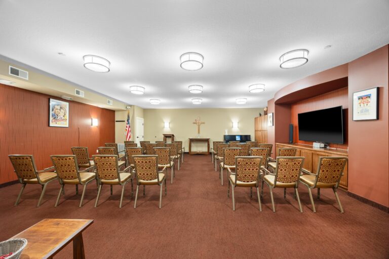 Chapel room with chairs facing an altar, cross on the wall, TV, and framed pictures on wood-paneled walls.
