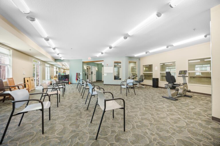 Room with spaced chairs, exercise equipment, and large windows, suggesting a physical therapy or rehab center.
