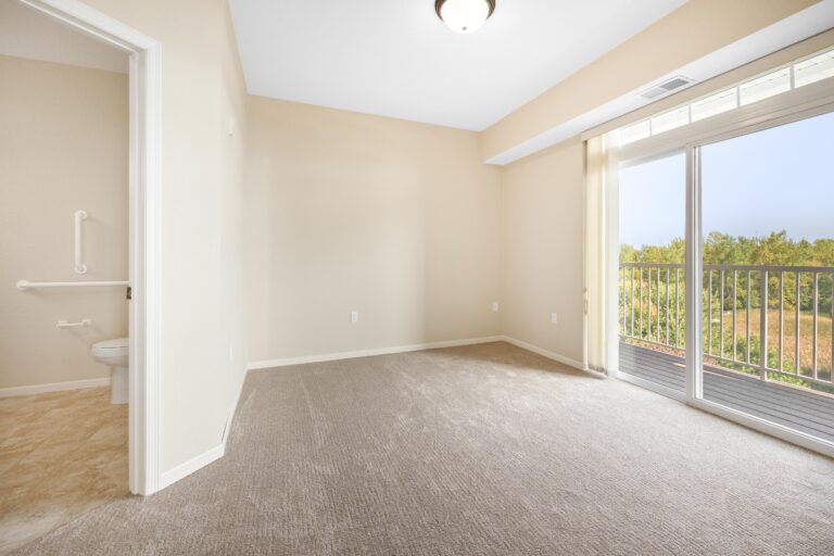 Empty carpeted room with sliding glass door to balcony; bathroom visible to the left; beige walls.