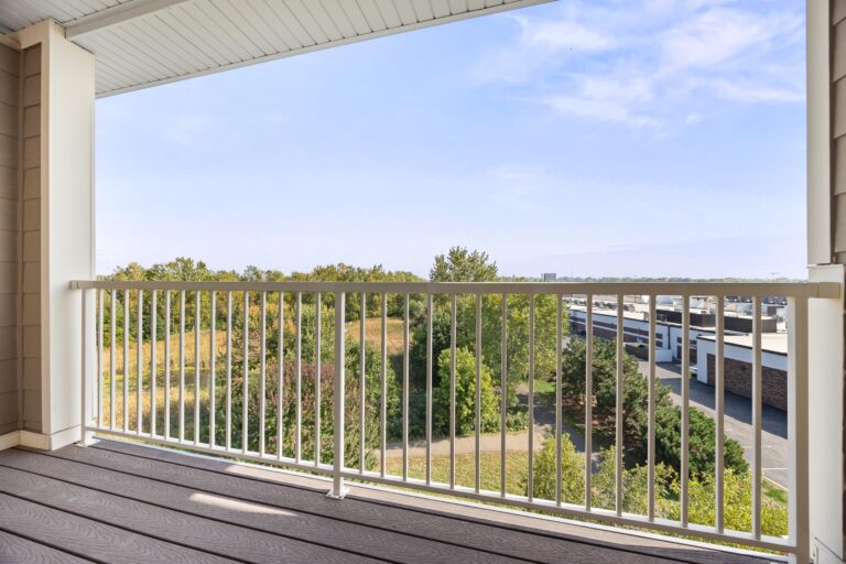 Second-floor balcony with white railing overlooking grassy area, trees, and distant buildings under a blue sky.