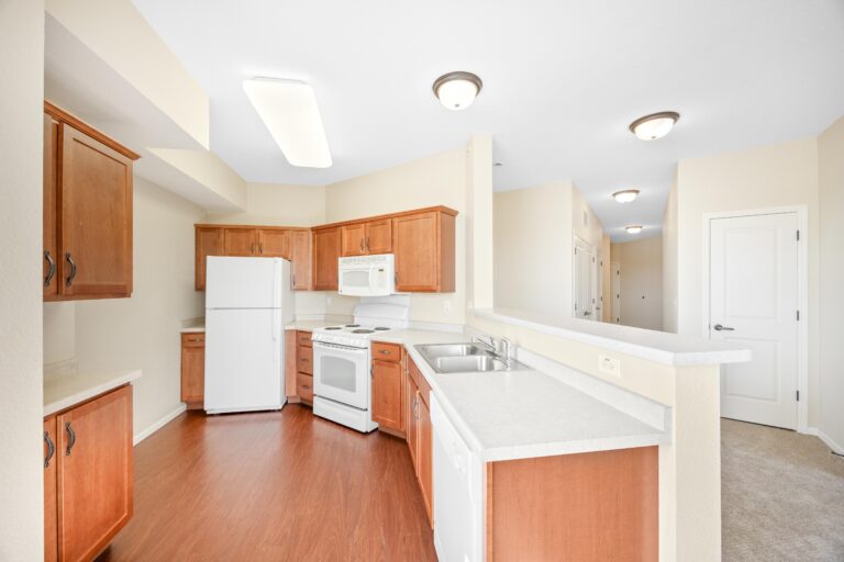 Bright kitchen with wooden cabinets, white appliances, and a double sink, next to a hallway and white door.