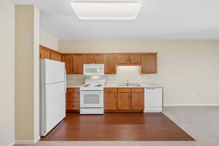 Small kitchen with wooden cabinets, white appliances, and a mix of hardwood and carpet flooring.