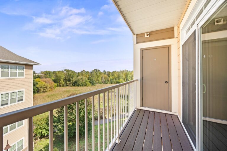 Modern apartment balcony with railing overlooking trees and a grassy area on a bright, clear day.