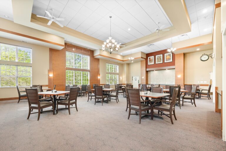 Spacious dining room with several tables and chairs, large windows, and chandeliers on a high ceiling.
