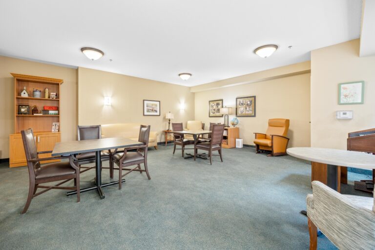 A spacious, well-lit room with tables, chairs, bookshelves, and artwork on beige walls.