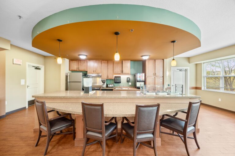 Modern communal kitchen with a curved countertop, pendant lights, and five chairs facing stainless appliances.