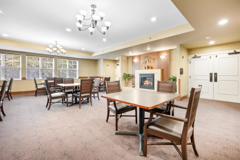 A spacious dining room with several tables, chairs, large windows, and a lit fireplace in the background.