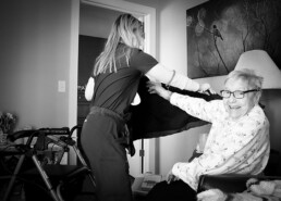 A caregiver helps a smiling elderly woman put on a sweater in a cozy room with a walker nearby.