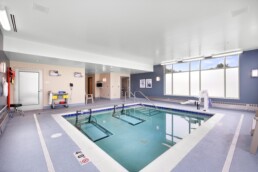 Indoor therapy pool with handrails, lift chair, and medical equipment beside large frosted windows.