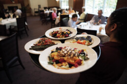 A server carries four plates of colorful meals in a busy restaurant dining room with people seated at tables.