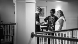 Three people stand talking indoors; one holds a notepad, while the others listen and smile.