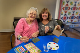 Two smiling women sitting at a table, painting and doing crafts together, with art supplies around them.