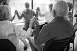 A group of elderly people sit in a circle, holding hands and smiling in a bright room.