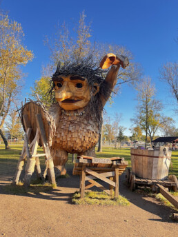Large wooden troll sculpture with raised arm, surrounded by autumn trees in a sunny park.