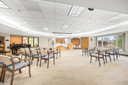 A bright chapel with rows of chairs, a piano, stained glass, and an altar at the front.