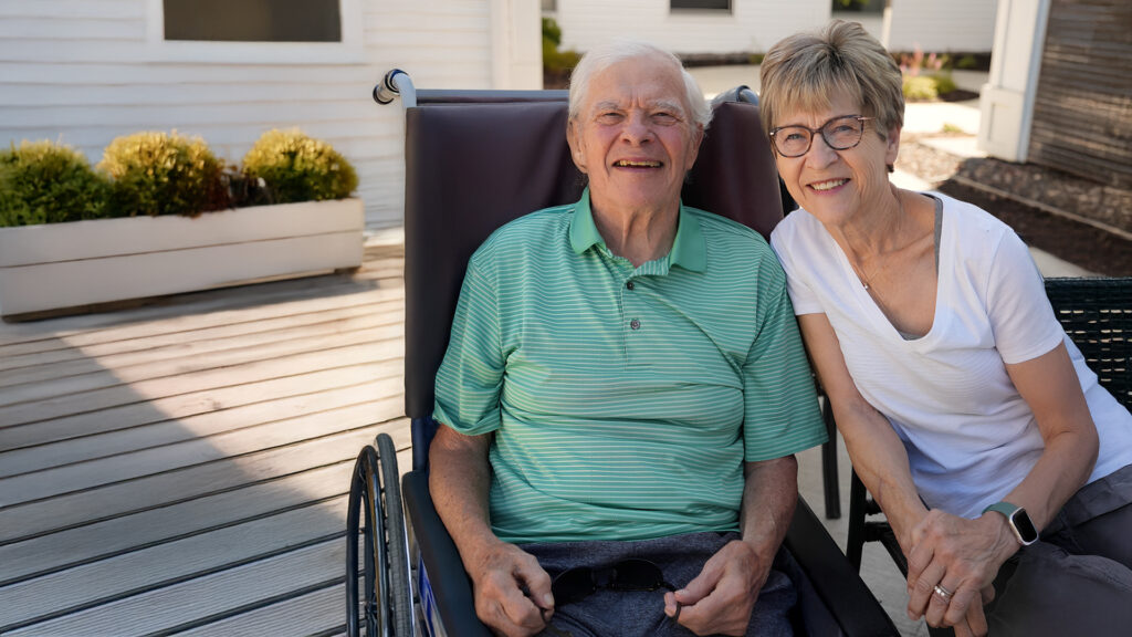 Senior man smiling in a wheelchair outside alongside an older woman smiling