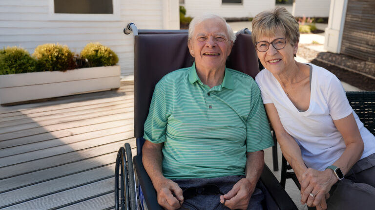 Senior man smiling in a wheelchair outside alongside an older woman smiling