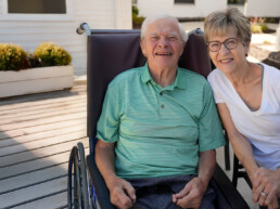 Senior man smiling in a wheelchair outside alongside an older woman smiling