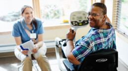 Smiling woman using exercise equipment, with a woman holding a clipboard nearby in a bright room.
