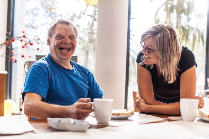 a man with Down Syndrome smiling and eating breakfast with a caregiver at an Adult Day Services center