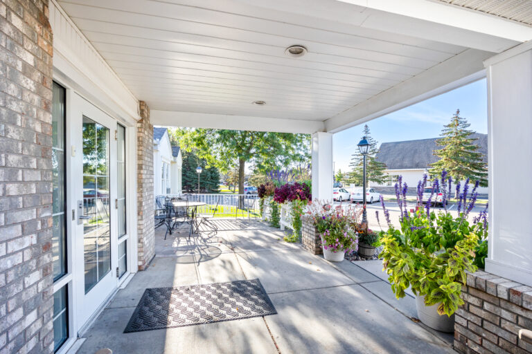 Covered patio with tables, chairs, potted plants, and a view of trees and parked cars in the background.