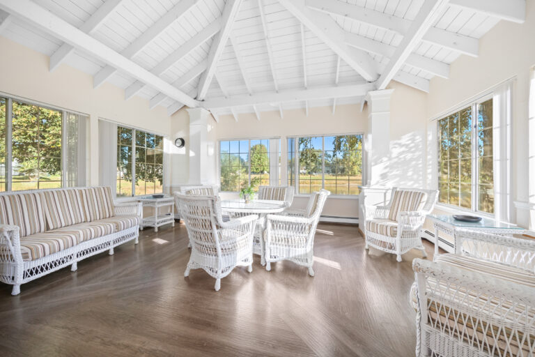 Bright sunroom with white wicker furniture, striped cushions, wood floors, and large windows overlooking greenery.