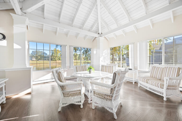 Sunlit room with white wicker furniture, striped cushions, and large windows overlooking green trees.