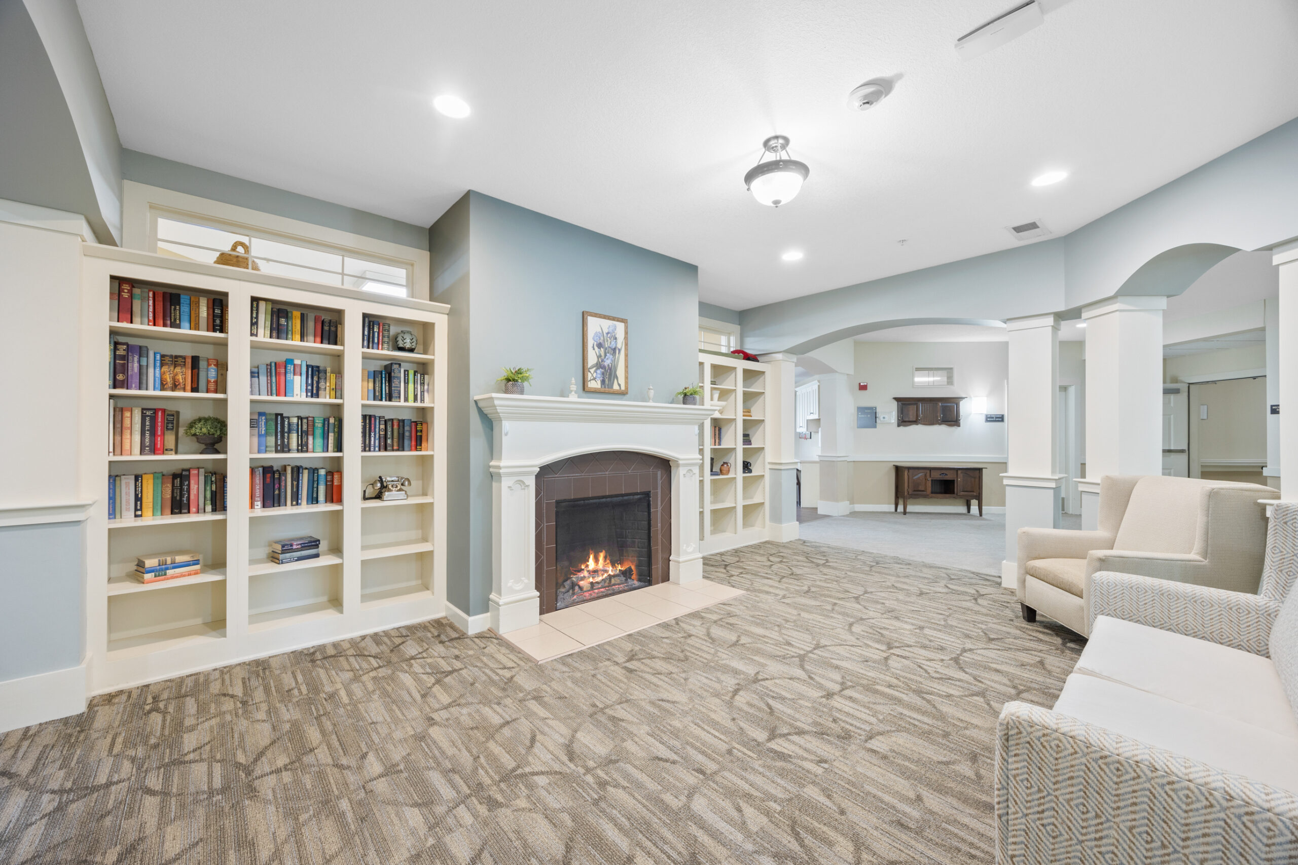 A cozy lounge with a fireplace, bookshelves, armchairs, and neutral decor in a well-lit room.