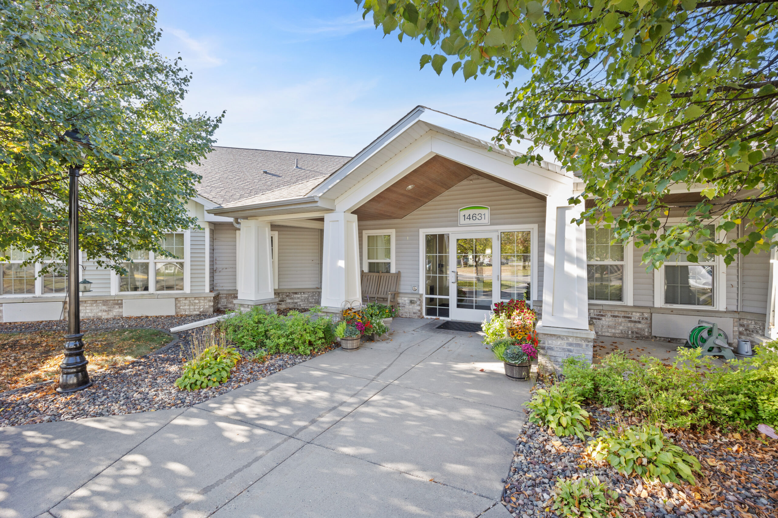 Single-story building entrance with large windows, a garden, and trees surrounding the concrete walkway.
