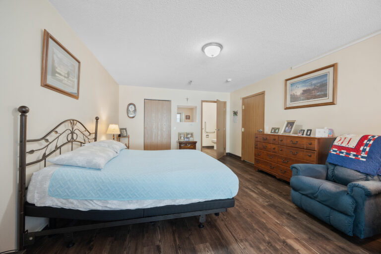 Bedroom with a metal bed, blue bedding, dresser, armchair, and wood flooring in a well-lit room.