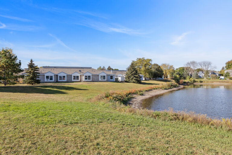Single-story white building beside a pond, surrounded by green grass and trees under a clear blue sky.