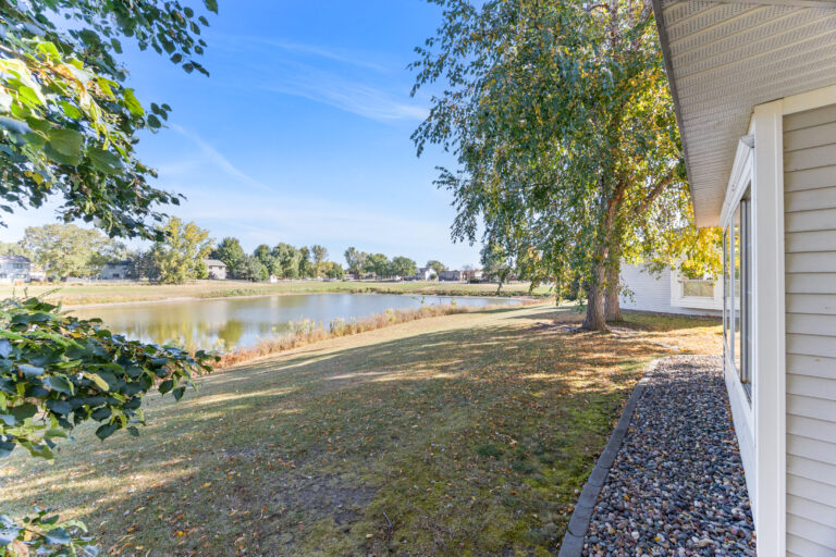 Lakeside backyard view with trees, grassy lawn, and clear blue sky next to a light-colored house.