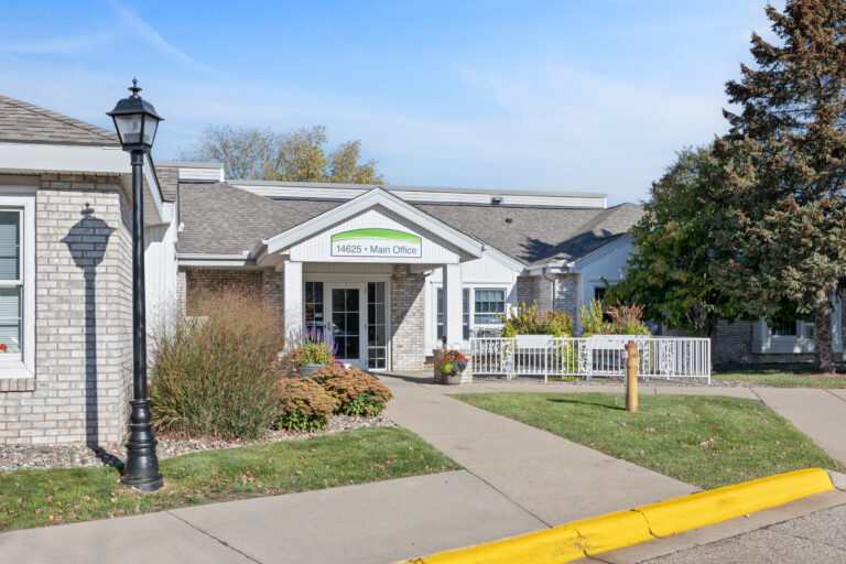 Single-story brick building with a Main Office sign, surrounded by plants and a sidewalk.