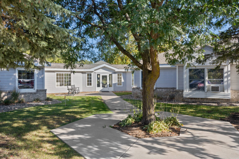 A sidewalk leads to a single-story house with blue siding, large windows, and a tree in front.