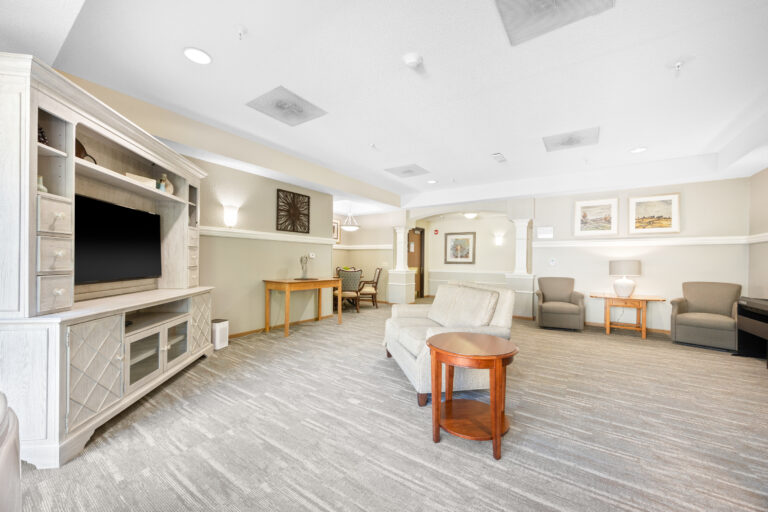 Bright, spacious lounge with chairs, tables, TV, and wall art in a neutral-toned, carpeted room.