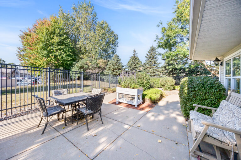 Sunny outdoor patio with a dining table, chairs, bench, and surrounding greenery next to a black fence.