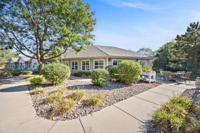 Single-story house with large windows, surrounded by shrubs, trees, and patio seating on a sunny day.