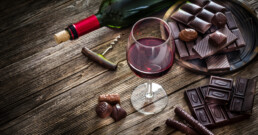 a rustic table with a plate of chocolates and a glass of red wine next to a wine bottle and cork screw.