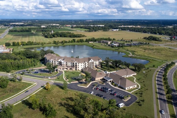 Aerial photo of Chateau Waters building and campus next to Lake Francis with water fountain feature in the middle of the lake and open fields stretching into the horizon to meet a partly cloudy blue sky.