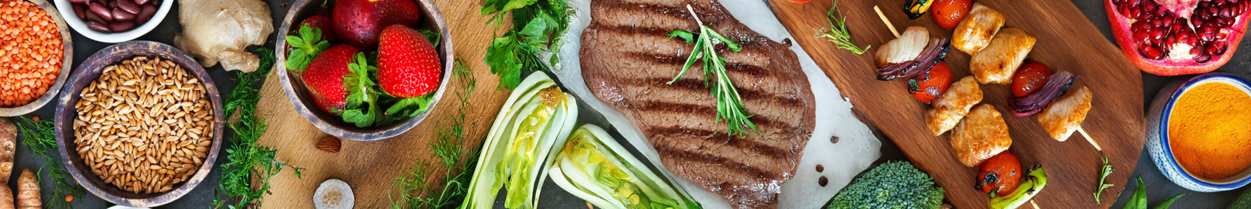 A variety of fresh foods arranged on a table, including a grilled steak, colorful vegetables, fruits, grains, and spices, creating a vibrant and appetizing display.