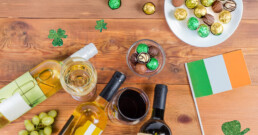 Bottles and glasses of wine, assorted chocolates, green shamrock decorations, grapes, and an Irish flag on a wooden table.