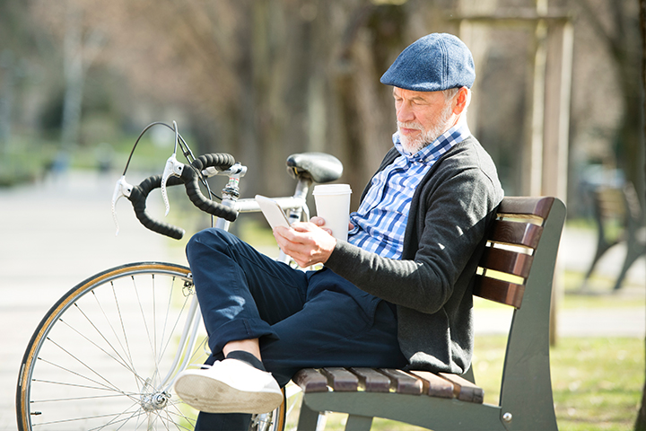 Older man sitting on a park bench with a coffee, looking at his phone; bicycle leaned beside him.