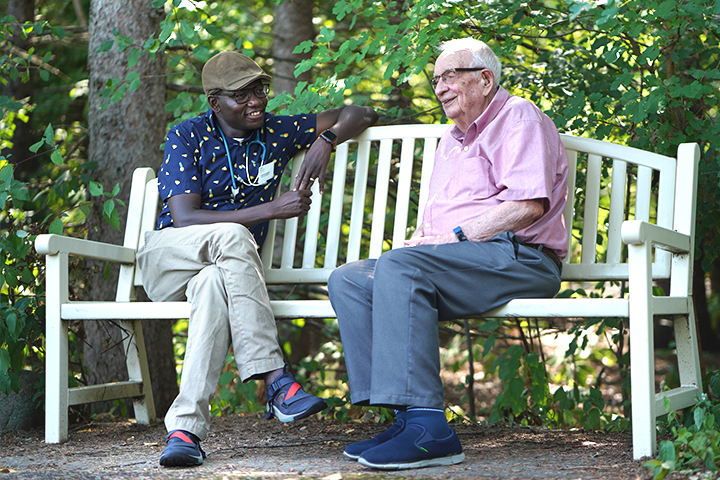 A male care team member engaged in discussion with an older male resident as they sit on a white park bench..