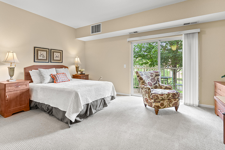 A bright and furnished bedroom with light-colored carpet and a sliding door leading to a private balcony.