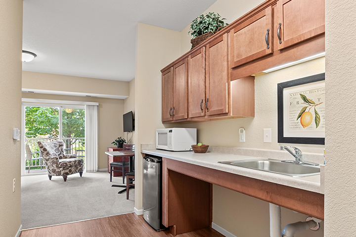 A kitchenette with small refrigerator, microwave, sink, and cabinetry with living space and sliding glass door with balcony in the background.