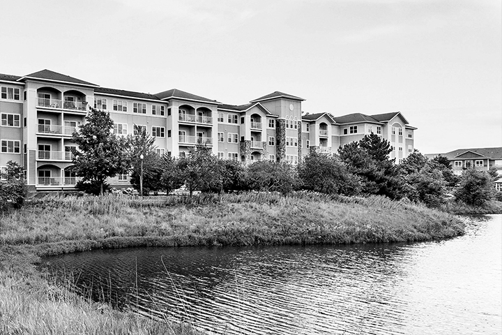 Black and white photo of a pond in the foreground with a grass-covered embankment lined by trees backdropped by a four-story building with lots of windows and balconies. A gazebo with green roof sits in the distance next to the pond.