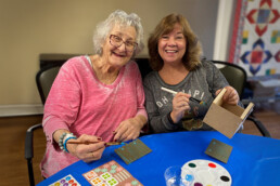 Two women smiling and enjoying a craft activity at a table with supplies and a paint palette.