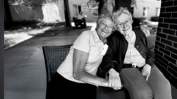 An older woman with her arm wrapped around another older woman. They are smiling. Black and white photo.